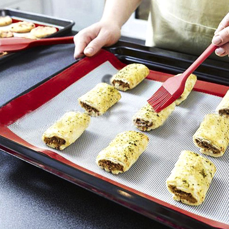 Person brushing pastry rolls with a red brush on a baking tray with silicone mat