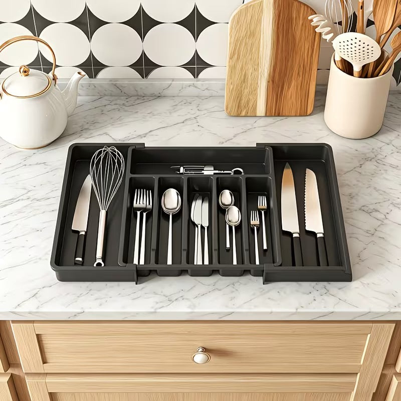 A black plastic kitchen utensil organizer tray holding stainless steel cutlery and knives, placed on a white marble countertop in a kitchen setting with a teapot, cutting boards, and utensil holder visible.