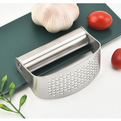 Metal garlic press on a cutting board with garlic, tomatoes, and a cookie.