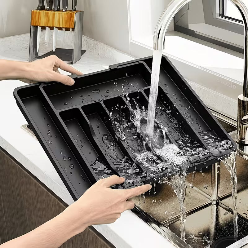 A black plastic kitchen sink organizer tray being rinsed under running water in a modern kitchen sink, with a person's hands holding the tray in place.